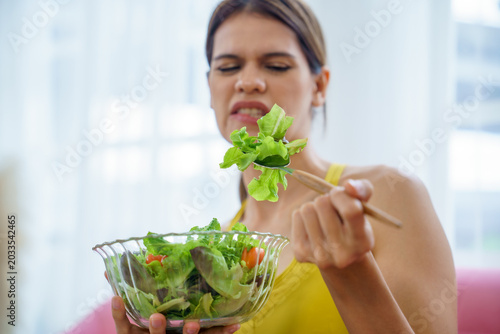 Woman salad lettuce bowl chopstick disgust grimace healthy diet indoor, young adult frowning at green leaf while holding glass bowl of mixed greens at home, picky eater
