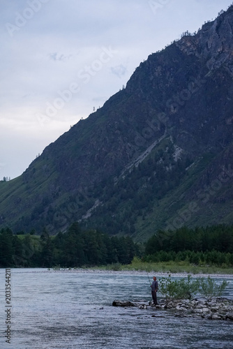 Fotografi A man fishes on a mountain river at the foot of a mountain in the evening