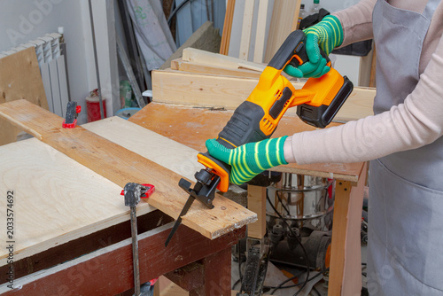 Foto Woman carpenter using cordless reciprocating saw to cut wooden board on workbench