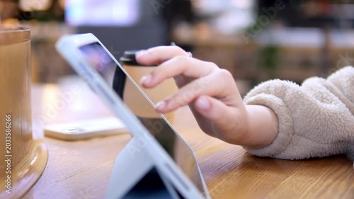 Close-up of child's hands using tablet at cafe table. Soft focus background shows cozy indoor setting. Child appears focused and engaged with device. Suitable for education, technology