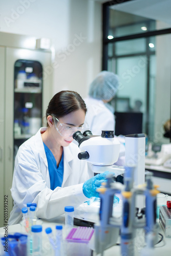 Young Asian scientist analyzing samples with microscope in a modern laboratory, focusing on medical research, biotechnology, healthcare, and innovation.