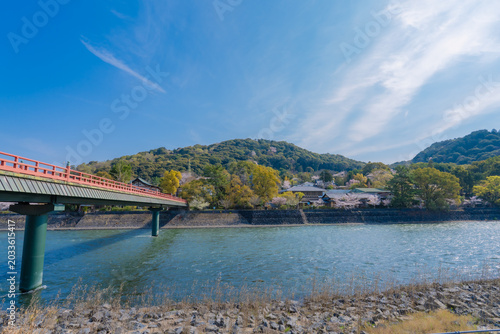Beautiful scenic view of Uji River flowing through Uji city in Kyoto, Japan, during spring season with blooming cherry blossom trees, surrounded by green mountains under a bright clear sky.