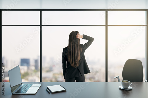 Canvastavla Businesswoman standing at office window in bright corporate workspace with city view, seen from behind, contemplating workday scene
