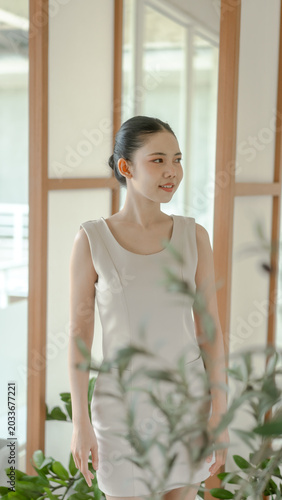 Young woman in a light sleeveless dress standing indoors near wooden-framed windows, soft natural light, green plants in foreground, calm and elegant portrait, neutral tones