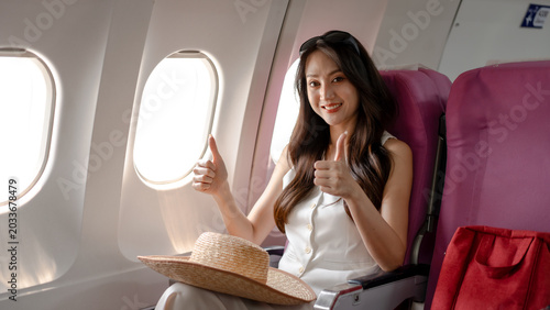 Fotografía Smiling woman passenger giving thumbs up while seated by airplane window in economy cabin, travel hat on lap and red bag nearby, bright daylight, vacation and air travel concept