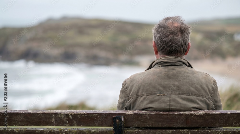 Fototapeta premium An older man sits on a wooden bench, looking out at a cloudy coastal landscape with a beach and hills in the distance.