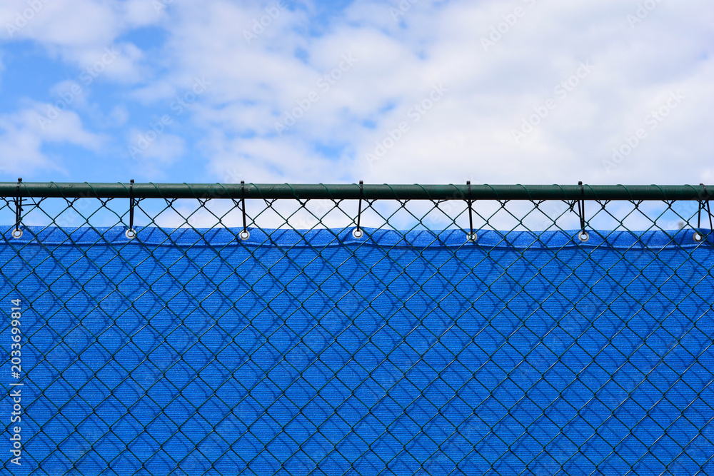 Fototapeta premium An eye level shot shows the top edge of a green wire mesh fence lined with a bright blue privacy screen against a partly cloudy sky.