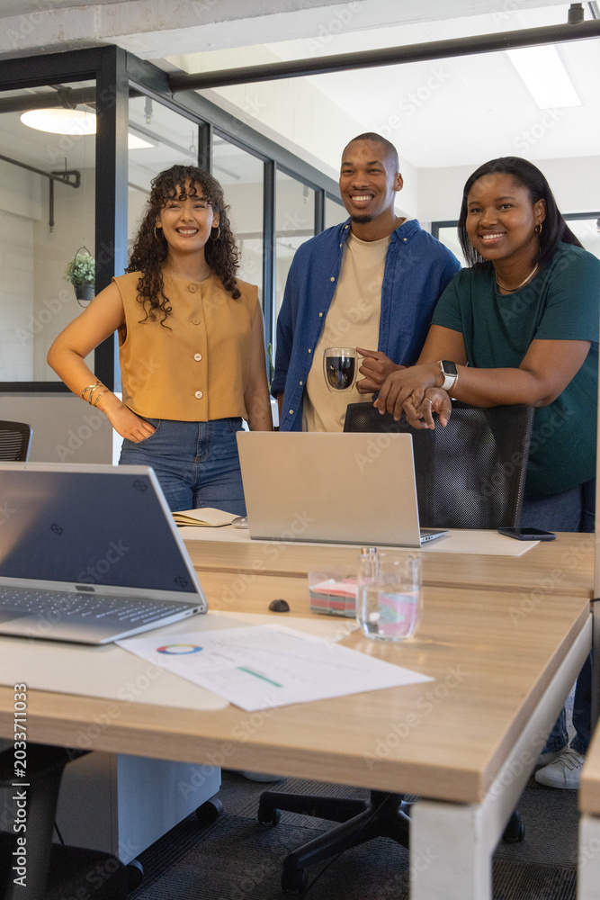 Fototapeta premium Diverse coworkers smiling and standing in modern office at wooden table with laptops, pie chart