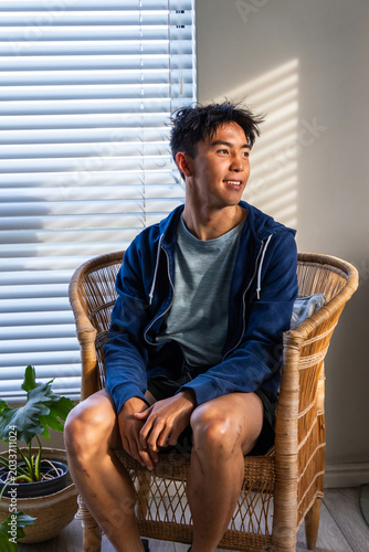 Bilde på lerret Asian male teen sitting at home in rattan chair by blinds, cushion, plant nearby