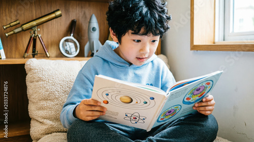 Young boy reading a book about space and science at home sitting on a couch