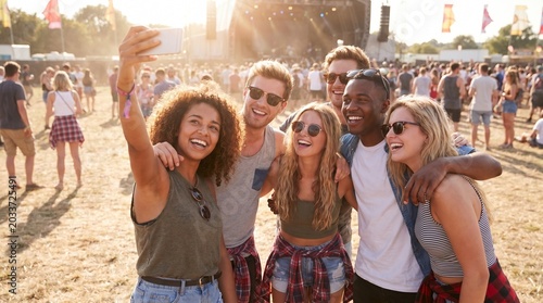 Diverse young friends taking a selfie, smiling and laughing. Enjoying an outdoor summer music festival with a large crowd and bright sunshine.
