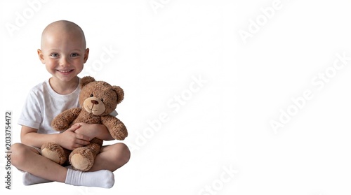 Smiling bald child wearing white t-shirt sitting cross legged holding brown teddy bear on white background, pediatric oncology patients, childhood cancer survival.