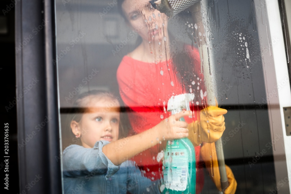 Fototapeta premium Woman and child cleaning a window with spray and squeegee indoors.