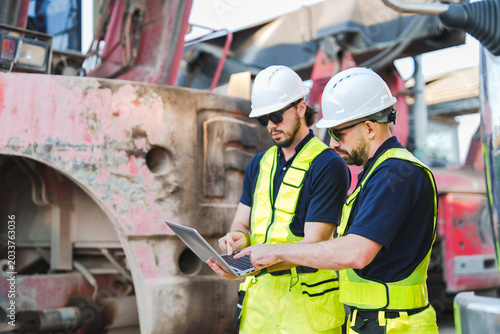 Construction worker using laptop industrial site inspection safety helmet reflective vest engineering team heavy machinery outdoor worksite equipment maintenance technical check teamwork