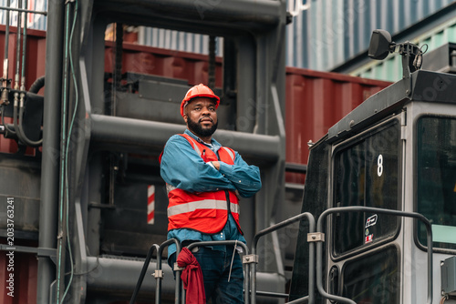 Port worker in hard hat and safety vest standing beside cargo crane at shipping terminal with arms crossed, focused and confident