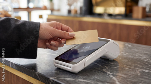 Close-up of a person holding a gold credit card to make a contactless payment on a wireless POS terminal at a cafe counter