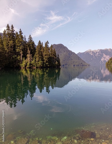 Serene Lake Reflection: Majestic mountains and lush evergreen trees form a stunning reflection on the glassy surface of a pristine lake, under a clear blue sky dotted with wispy clouds.