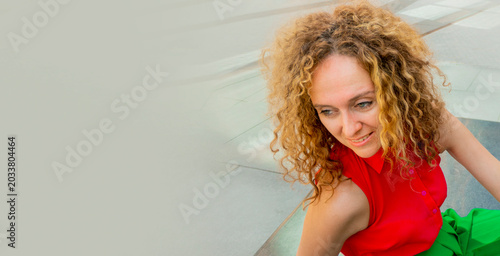 Fotografia Fashionable woman in bright clothes, with long blond curly hair, is sitting in the lotus position on the street basking in the sun and resting against the background of a modern business center