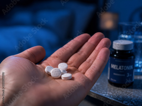 A person's hand holds white melatonin pills, with a blurred bottle and glass of water on a bedside table in a dimly lit bedroom at night, illustrating a routine for improving sleep, promoting relaxati