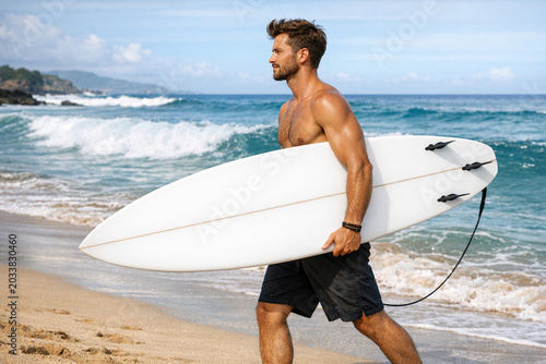 A young male surfer holding a white surfboard while walking towards the ocean. He is shirtless and wearing black swim trunks.