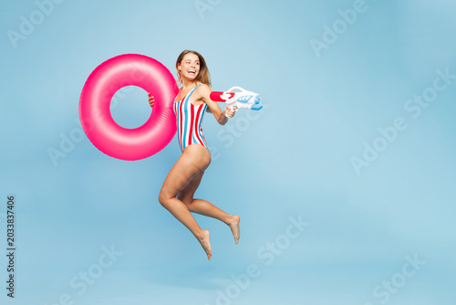 Full body young fun woman she wear bikini swimsuit rest near hotel pool hold pink rubber ring play shoot from water gun jump high isolated on plain blue background Summer vacation sea sun tan concept