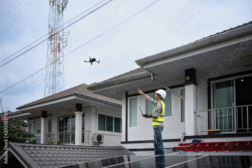 Asian male engineer in safety vest and hard hat operates a drone while holding a laptop to inspect solar panels on a house rooftop, ideal for renewable energy and technology concepts