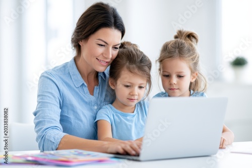 Woman teaching two young girls online using a laptop at a bright, modern home workspace, with colorful educational materials and a plant visible in the background