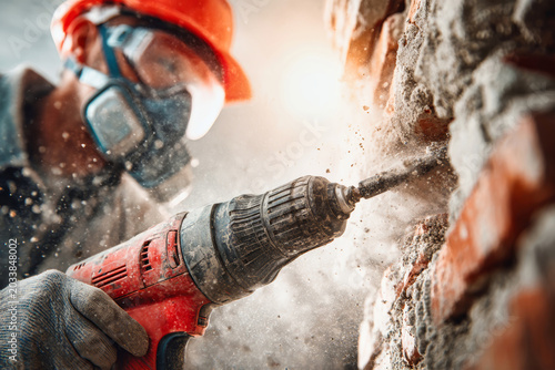 A construction worker operates an electric drill on a brick wall while dust fills the air in a well-lit space. The worker wears a helmet and mask
