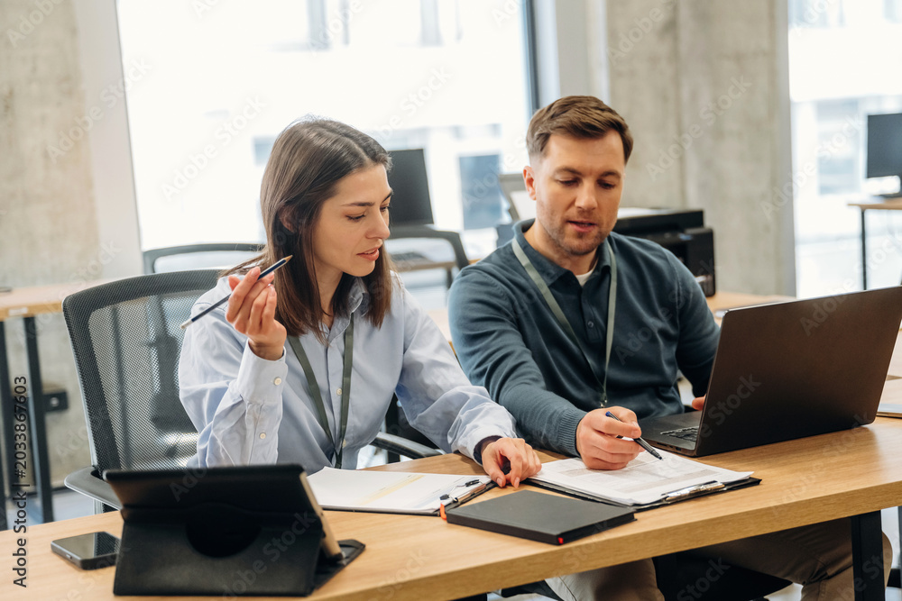 Fototapeta premium Two professionals engaged in discussion at a modern office workspace, one woman holding a pen and the man pointing at a laptop screen with documents on the table