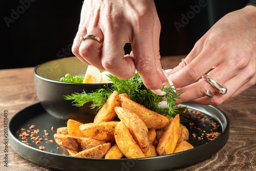 Schilderij op canvas Hands of a chef garnishing crispy fried potato wedges with fresh dill and hard-boiled egg on a black plate, close-up view