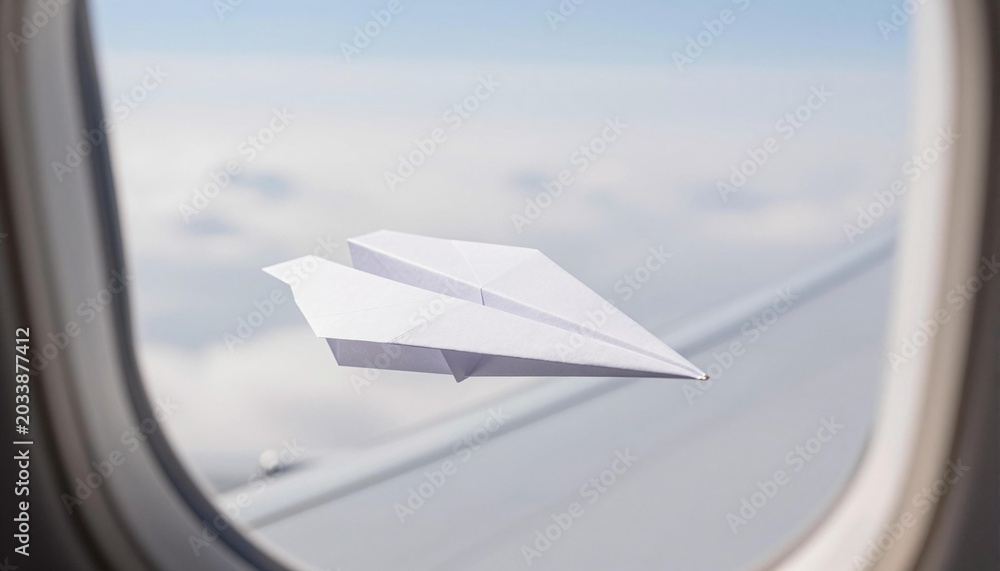 Fototapeta premium White paper airplane flying past an airplane window over a cloud layer with the wing and soft sky visible