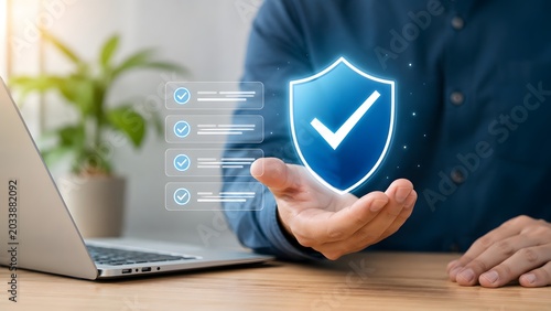 Hand presenting glowing digital shield and checklist hologram over laptop for cyber security assurance and software validation