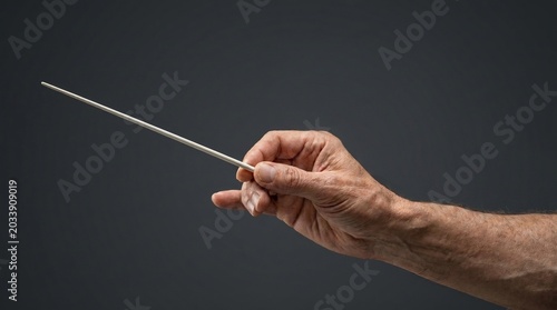 Close-up of a wrinkled elderly hand holding a thin white conductor baton on a dark background