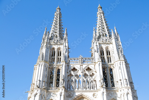 A detailed view of the towers of Burgos Cathedral, showcasing its Gothic spires, ornate stone carvings, and pointed arches against a bright blue sky.