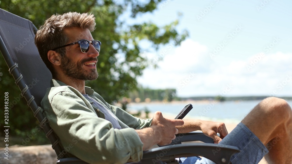 Obraz premium A man is sitting in a chair by the water, smiling and holding a cell phone