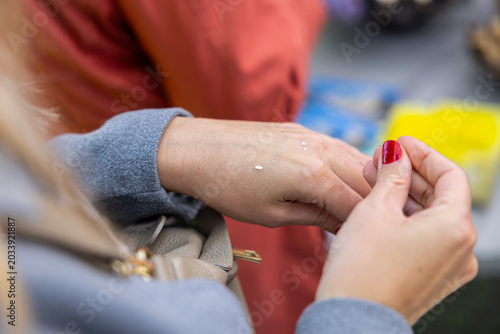 Close-up of hands applying small dots of cream on a wrist during product sampling, with red manicure and blurred market background