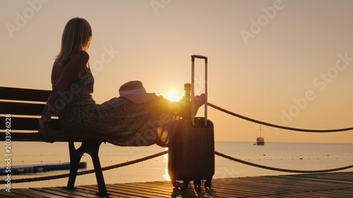 Woman sitting on bench by sea at sunrise with suitcase and hat, peaceful travel contemplation.