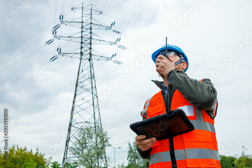 Technician or inspector evaluating electrical grid infrastructure damage prevention during a pre-storm site survey. Energy engineer speaks on portable radio for real time reporting. 