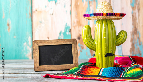 Fiesta Fun Cactus Decoration: A vibrant, green cactus adorned with a traditional sombrero and a playful mustache stands proudly on a colorful serape, next to a rustic wooden frame.