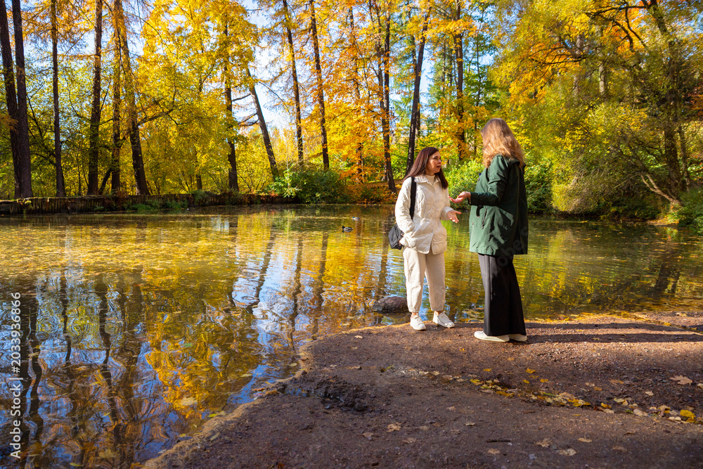 Fototapeta premium Two woman talking in autumn park near pond. Friends walking outdoors on bright fall day. Conversation in nature, leisure time lifestyle, seasonal recreational activity, community.