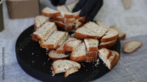 Close-up of sliced bread pieces on a black serving tray, with a gloved hand cutting fresh loaf on a patterned tablecloth