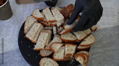 Close-up of sliced bread pieces on a black serving tray, with a gloved hand cutting fresh loaf on a patterned tablecloth