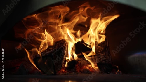 Close-up of burning logs and bright flames inside a wood-fired street food pizza oven, with warm glow, charred texture, and dark copy space