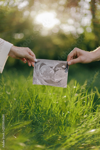 Couple holding ultrasound scan image of baby outdoors in green field.