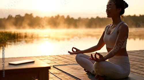 Asian woman meditating on wooden pier at misty sunrise lake