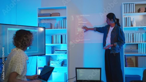 A professional male software developer stands presenting a data script and system infrastructure to his colleague during a late-night tech briefing, looking at camera while lines of code are projected