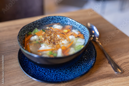 Hearty soup in blue speckled bowl. Carrots, celery, and dumplings float inside. Golden topping adds crunch and color. Wooden table and spoon complete the scene. Natural lighting highlights texture