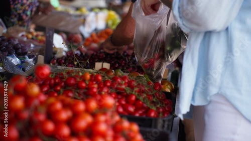 Customer selecting fresh organic produce at vibrant outdoor farmers market, placing ripe cherry tomatoes and sweet cherries into shopping bag for healthy eating and local food lifestyle.