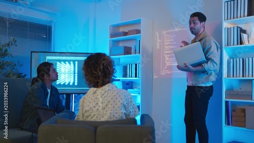 A professional male software engineer stands presenting and explaining lines of code projected on a wall display during a technical meeting, gesturing towards the data script in a dark room with drama