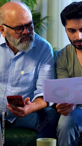 Indian retired father and young son working together on laptop and smartphone at home, reviewing financial report, share trading data and online business documents while discussing profit and loss